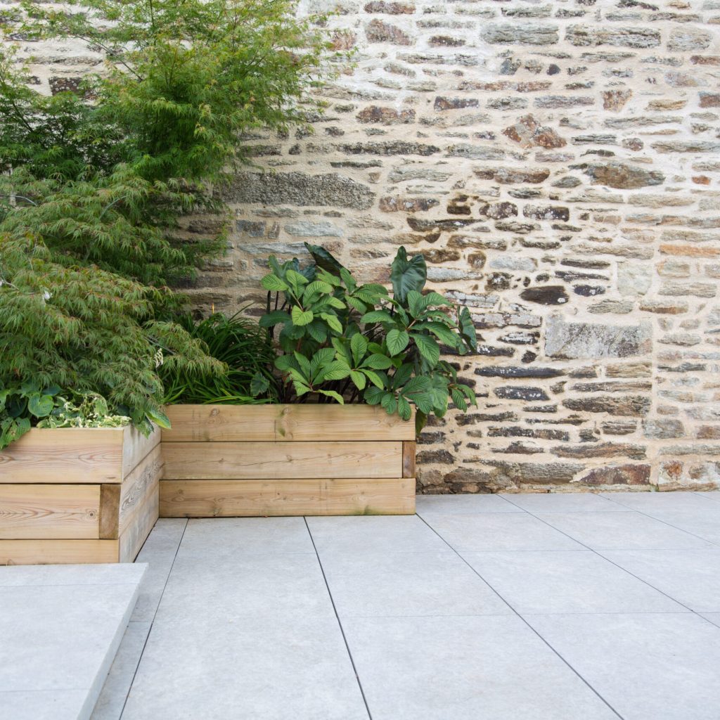 Modern garden design and terrace construction: Hillside plot paved with natural sidewalk flagstones in contrast to raised beds planted with green plants in wooden pots in front of an old stone wall